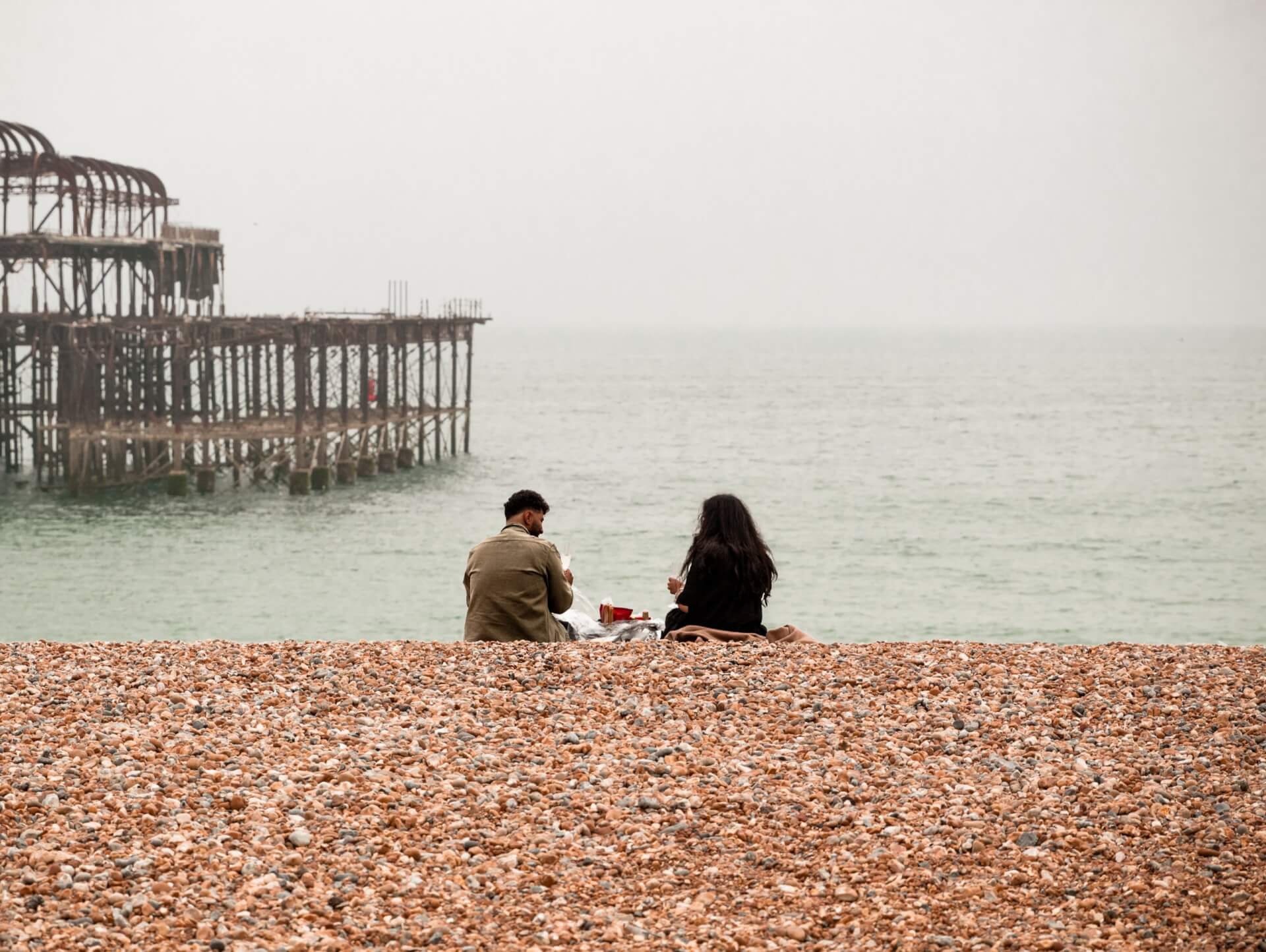 Two individuals relax on the beach, gazing at the historic West Pier in the background.