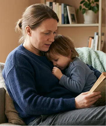 A mother sits on a couch, reading a book to her young child, who is attentively listening and looking at the pages.