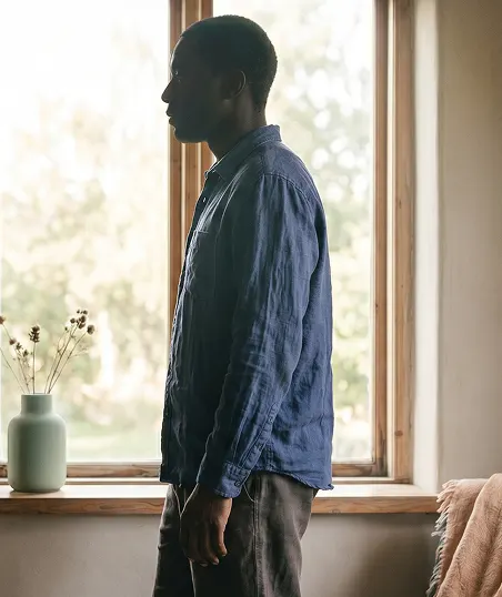 A man stands by a window, looking outside, with a thoughtful expression as natural light streams in.