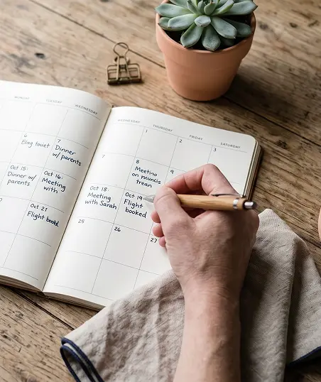 A close-up of a person's hand writing notes in a planner with a pen on a wooden table.