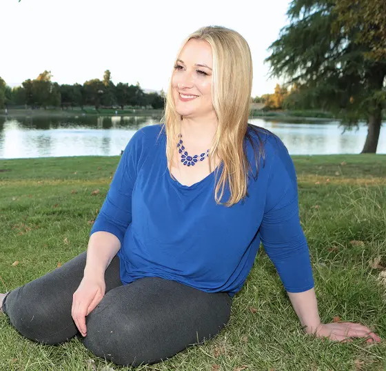 Kristin Carl wearing a blue shirt and necklace sits on the grass, enjoying a sunny day outdoors.