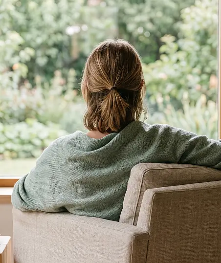 A woman sits on a couch, gazing thoughtfully out of a window, with natural light illuminating the room.