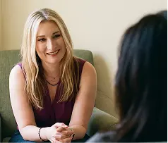 A woman seated in a chair engages in conversation with another woman nearby.